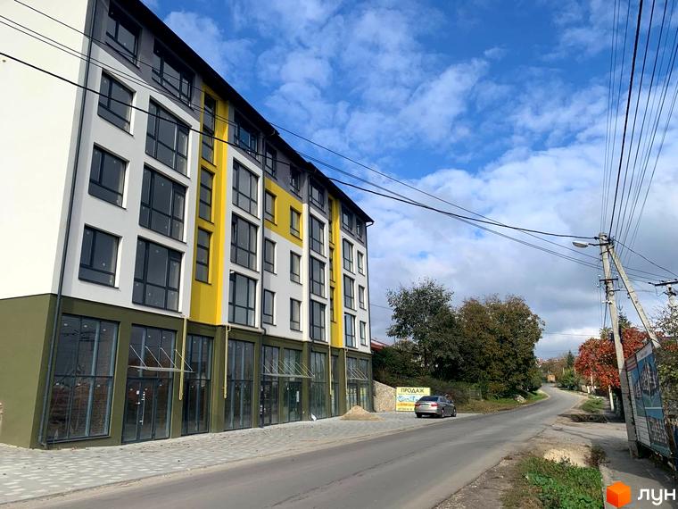 Багатоповерховий будинок Житловий комплекс ПАНОРАМА з великими вікнами та скляними вхідними групами. Навколо будинку асфальтована дорога та зелені насадження.