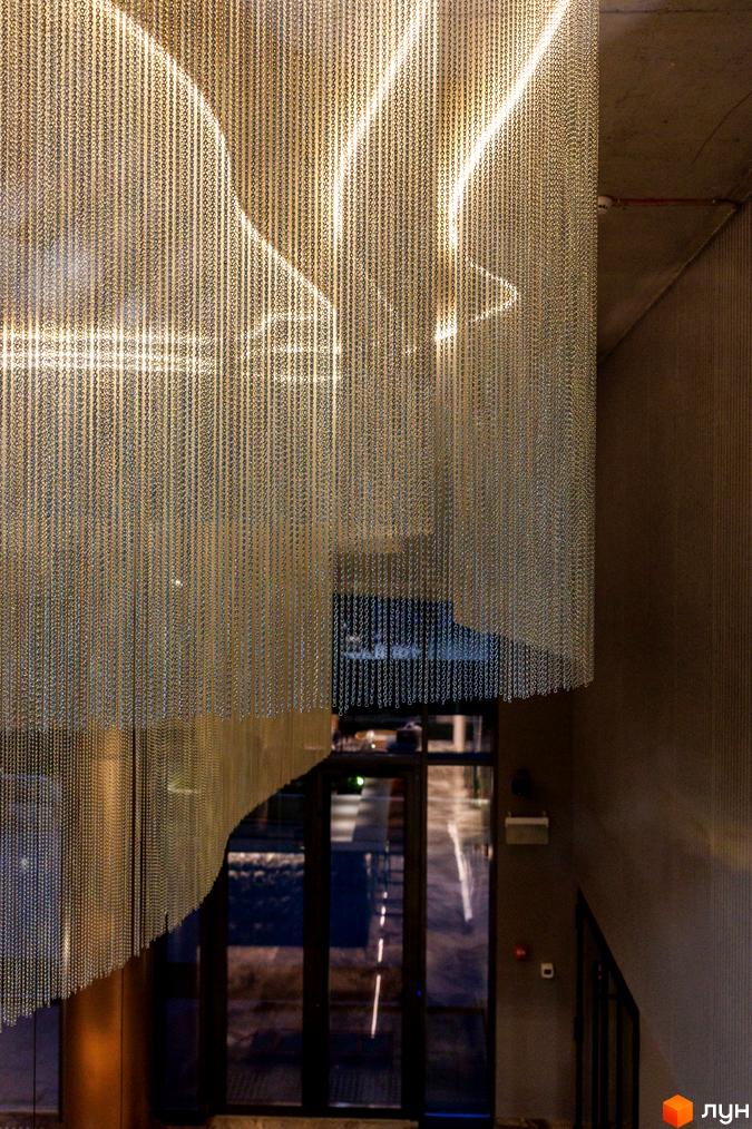 Житловий комплекс Avalon Prime з унікальним дизайном фасаду з металевих ланцюгів, що створює гру світла та тіні. Вхідна група з скляними дверима та сучасним освітленням.