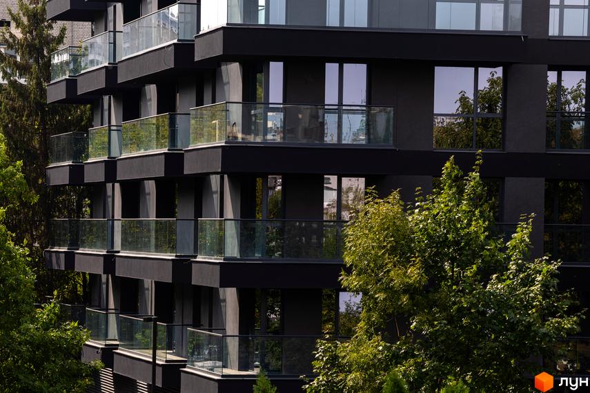 Житловий комплекс Avalon Prime з темним фасадом та скляними балконами, оточений зеленню. Зовнішній вигляд новобудови з численними вікнами та виступаючими елементами.
