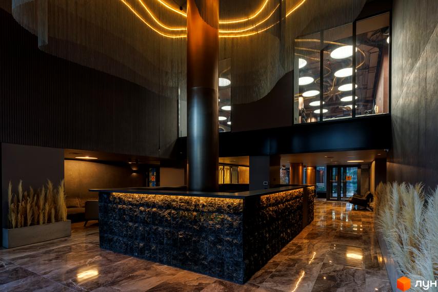 Інтер'єр лобі житлового комплексу Avalon Prime з великою колоною та підсвіченою стійкою ресепшн. Сучасний дизайн з натуральними матеріалами та декоративним освітленням.