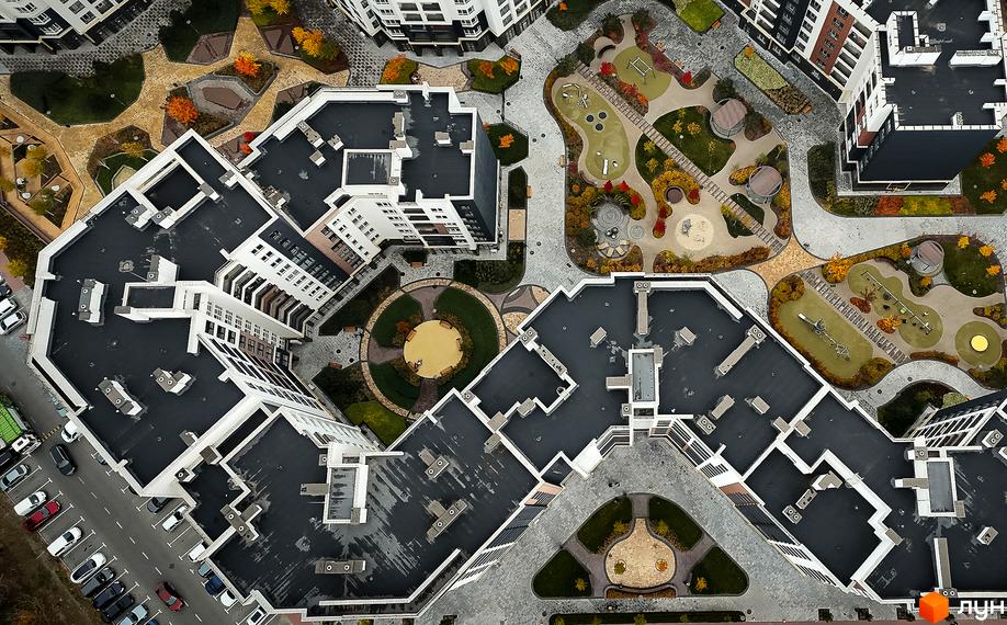 Вид зверху на новобудову ЖК Синергія Сіті з білими фасадами та темними дахами, що оточена упорядкованою прибудинковою територією з дитячими майданчиками та зеленими зонами.