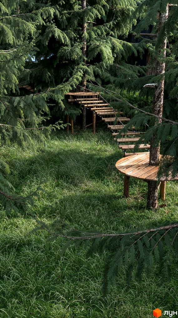 Прибудинкова територія житлового комплексу Guta Nova з зеленим газоном та дерев'яними сходами серед хвойних дерев.