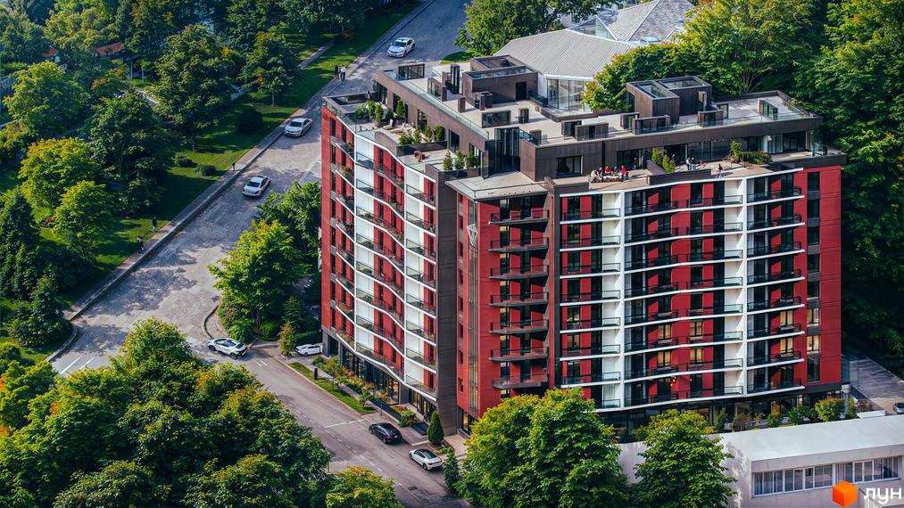 Житловий комплекс Рубін Апартамент з червоно-білим фасадом та балконами, оточений деревами. Поруч дорога з автомобілями та зелена зона.