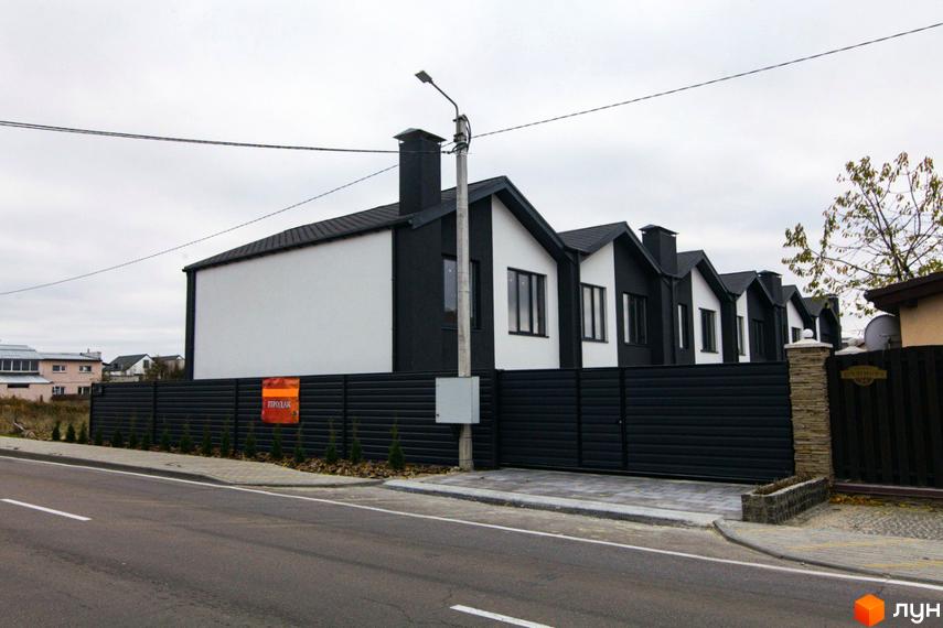 Зовнішній вигляд новобудови на вул. Шалімова, 36. Фасад будинку чорно-білий, з темною черепицею. Поруч огороджена територія з бруківкою та зеленими насадженнями.