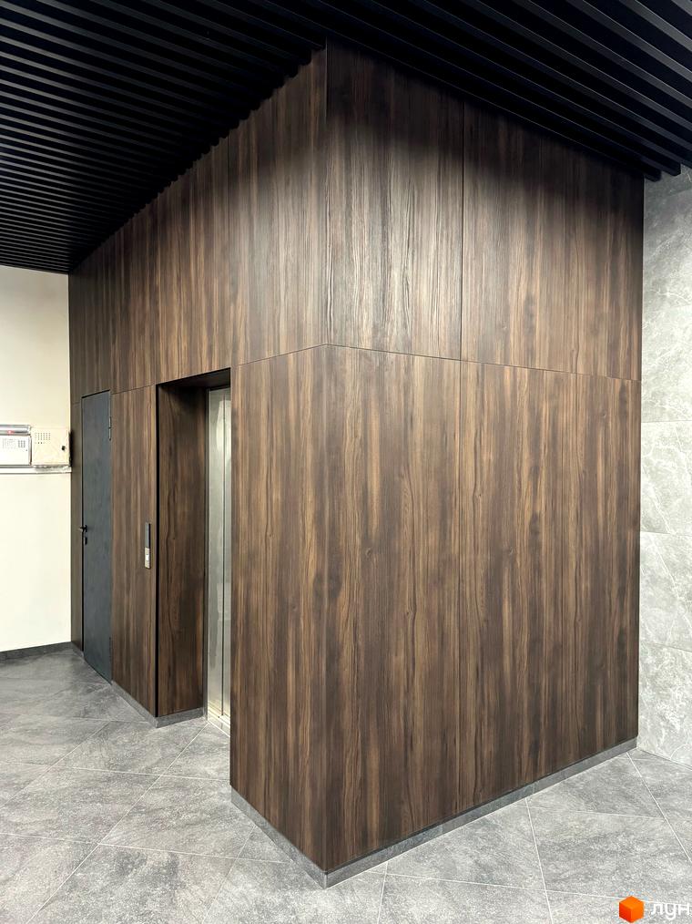 Сучасний дизайн інтер'єру з дерев'яними панелями та сірою плиткою біля ліфта в житловому комплексі GREENHOUSE CITY. Оздоблення стін темним деревом створює затишну атмосферу.