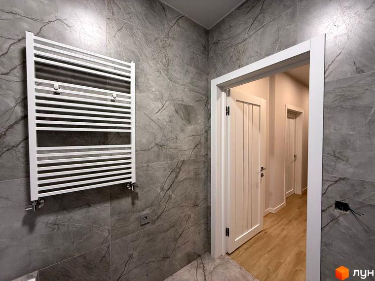 Дизайн ванної кімнати з сірим мармуровим оздобленням та білим рушникосушаркою. Відкриті двері ведуть до коридору з дерев'яною підлогою.