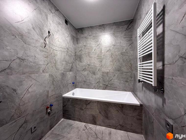 Ванна кімната в ЖК Comfort City з сірою плиткою під мармур та білою ванною. Поруч розташований білий рушникосушарка.