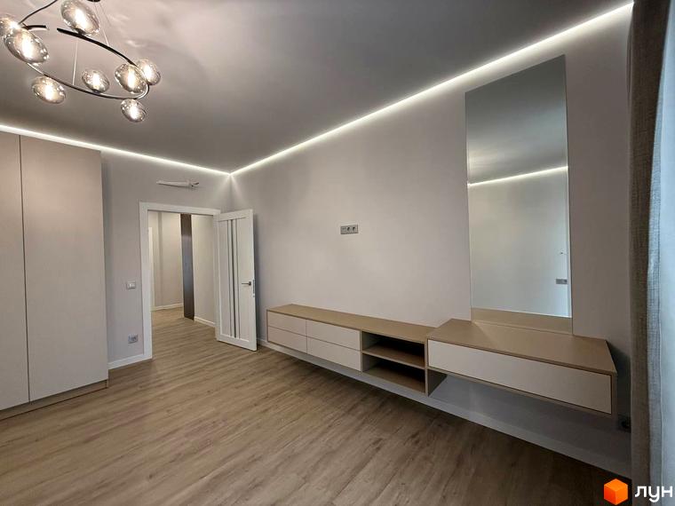 Дизайн спальні в ЖК Comfort City з підсвічуванням стелі та великим дзеркалом. Простора кімната з підлогою під дерево та вбудованими меблями.