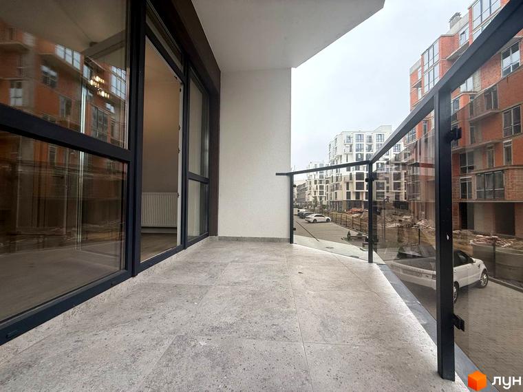 Балкон з скляною огорожею та видом на цегляні та білі багатоповерхові будинки ЖК Comfort City. На території комплексу видно припарковані автомобілі та дорогу.