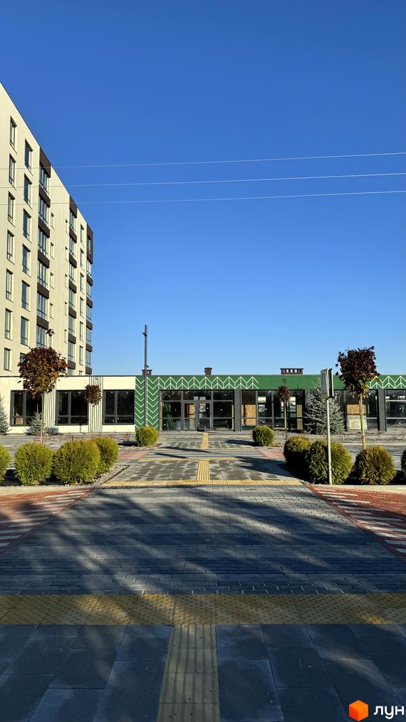 Житловий комплекс GREENHOUSE CITY з багатоповерховим будинком та одноповерховою будівлею з зеленим фасадом. Прибудинкова територія з бруківкою та кущами.