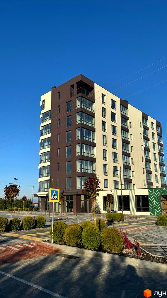 Новобудова ЖК GREENHOUSE CITY з біло-коричневим фасадом та великими вікнами. Поруч розташована прибудинкова територія з кущами та пішохідним переходом.