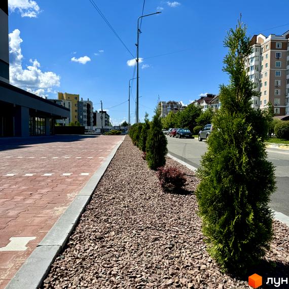 Зовнішній вигляд житлового комплексу Idilika Avenue з червоною бруківкою та зеленими насадженнями вздовж дороги. Навколо житлові будинки та міська інфраструктура.