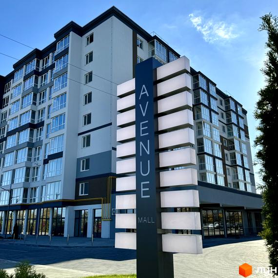 Зовнішній вигляд новобудови Idilika Avenue з фасадом у сірих та білих тонах та скляними балконами. Перед будинком розташована стела з назвою комплексу та освітленням.