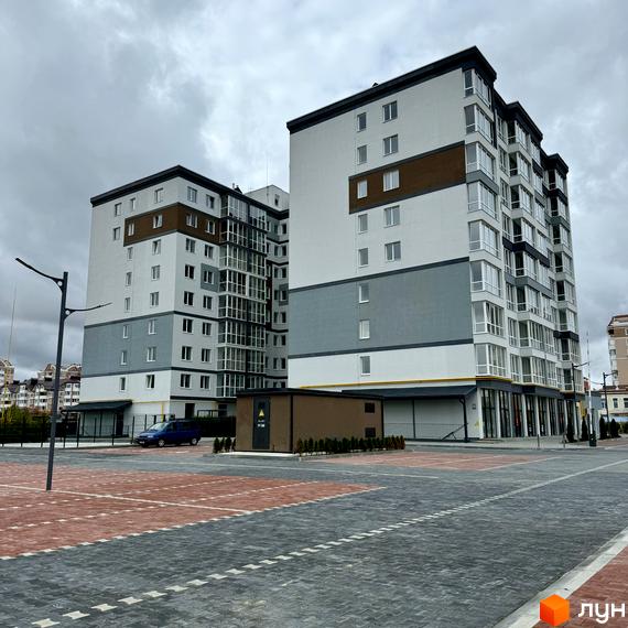 Новобудова ЖК Idilika Avenue з сірими та білими фасадами, коричневими вставками та скляними балконами. Поруч розташований паркінг та зелені насадження.