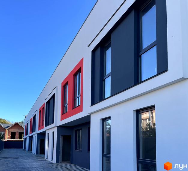 Ряд таунхаусів Belissimo з білими фасадами, темно-сірими елементами та червоними віконними рамами. Перед будинками вимощена доріжка.