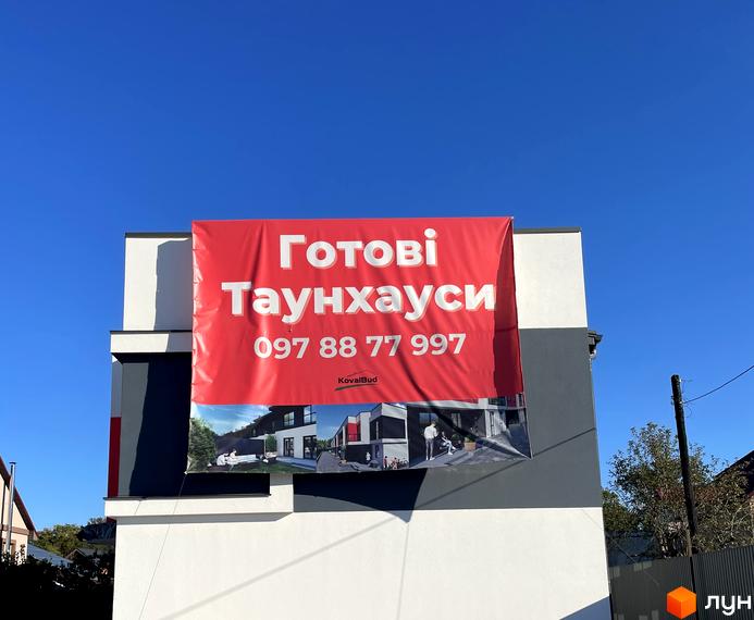 Зображення готового таунхаусу Belissimo з білими та темно-сірими фасадами. На будівлі розміщено банер з інформацією про продаж таунхаусів.