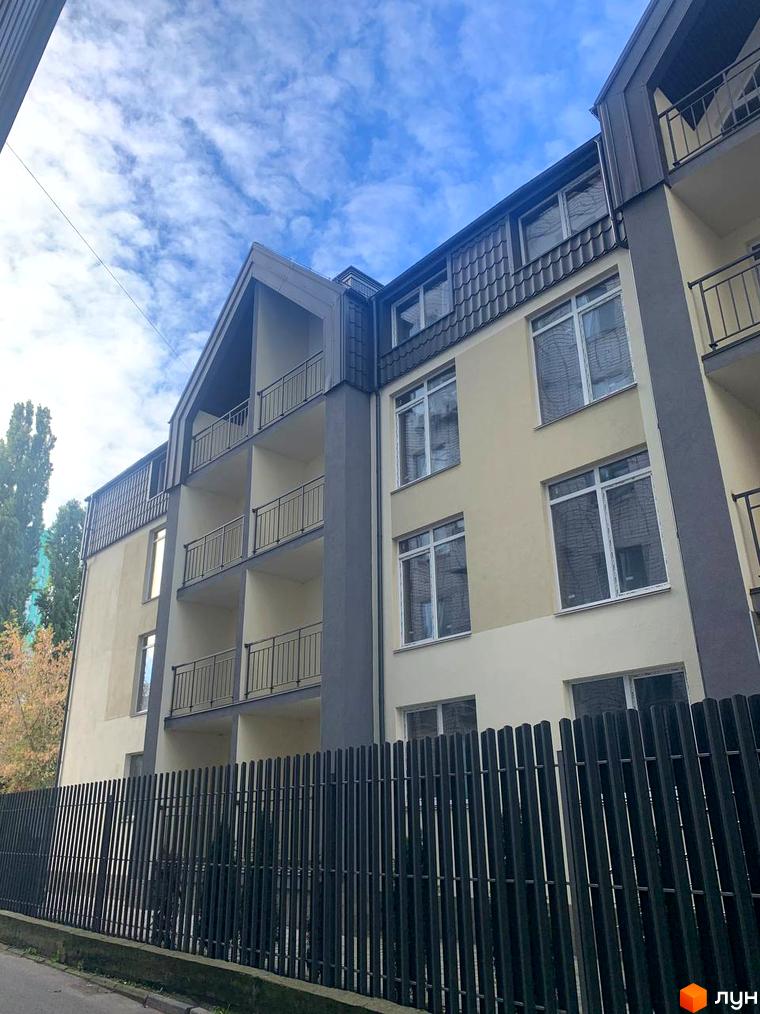 Клубний будинок „На Макіївській“ з балконами та великими вікнами, огороджений парканом. Навколо будинку зелені насадження та блакитне небо з хмарами.