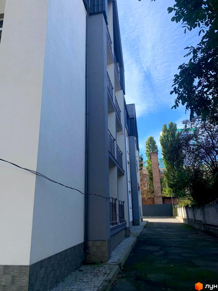 Клубний будинок „На Макіївській“ з сірими та білими фасадами, балконами з чорними поручнями та бруківкою біля входу. Поруч зелені дерева та блакитне небо.