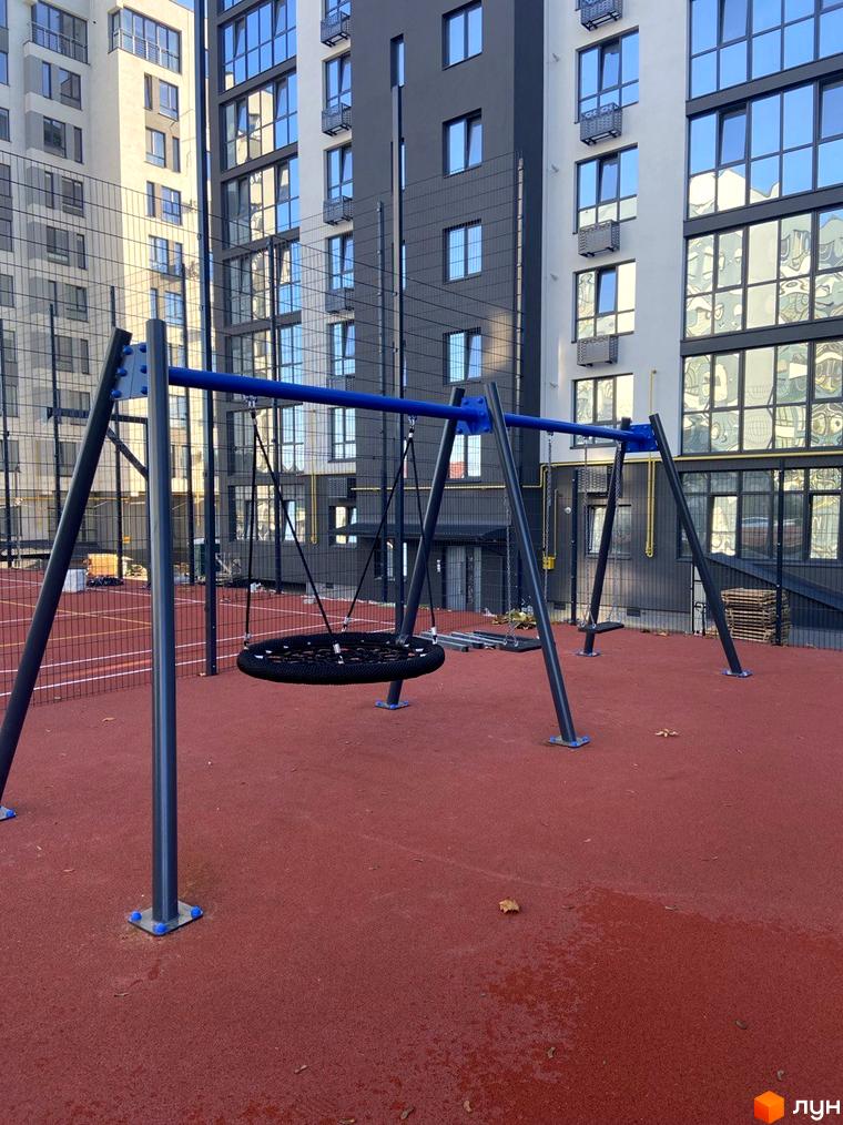 Дитячий майданчик з гойдалкою-гніздом біля багатоповерхових будинків ЖК Comfort House. Територія комплексу має червоне покриття та огорожу.