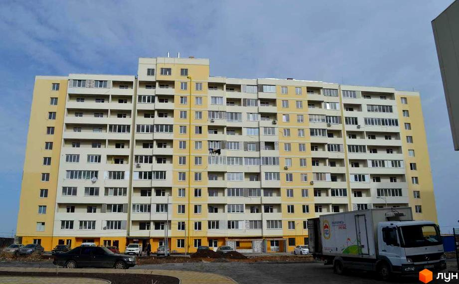 Висотна будівля житлового комплексу з жовтими та білими фасадами. На передньому плані прибудинкова територія з автомобілями та вантажівкою.