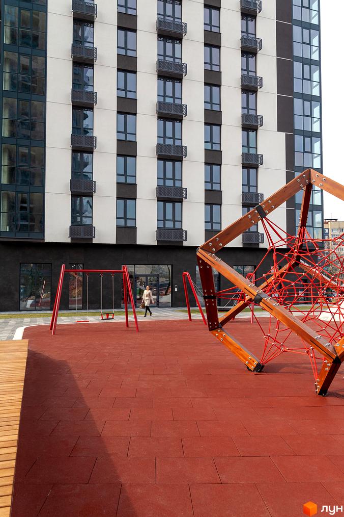 Житловий комплекс Avalon Up з фасадом з білої та темної плитки, великими вікнами та мініатюрними балконами. На передньому плані дитячий майданчик з червоним покриттям та ігровими елементами.