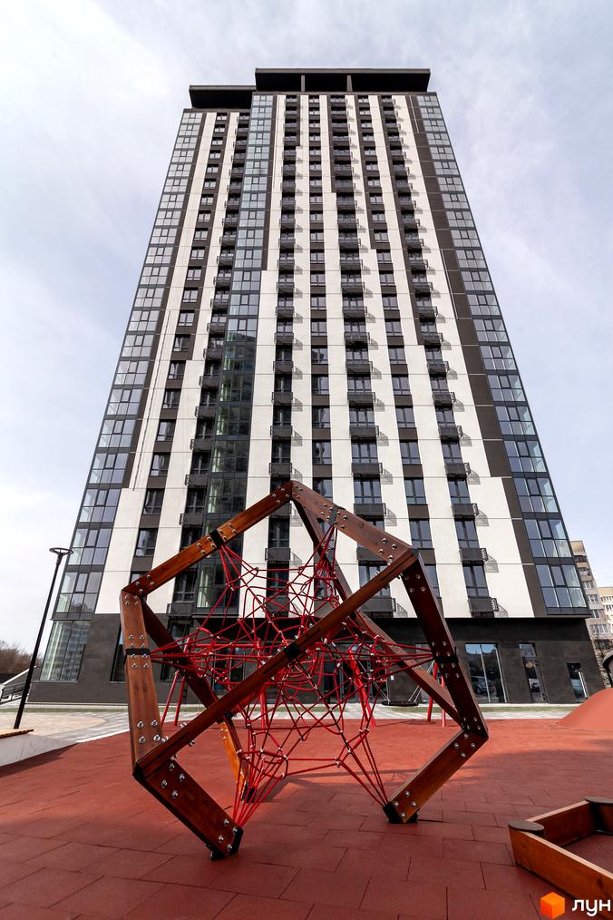 Житловий комплекс Avalon Up з фасадом з білих та темних смуг, вікнами та балконами. На передньому плані дитячий майданчик з дерев'яною конструкцією та червоною сіткою.