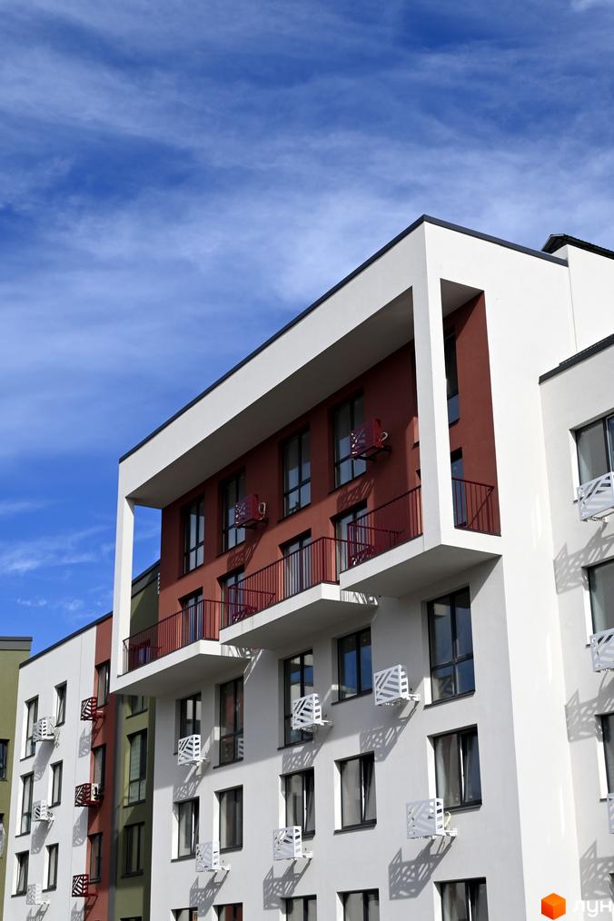 Будівельний проєкт ЖК Субурбія. Фасад будинку з червоними та білими елементами, балконами та кондиціонерами на фоні блакитного неба.