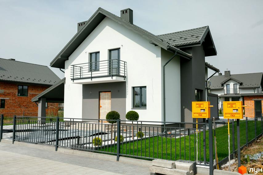 Котеджне містечко KM Praud Hills: двоповерховий будинок з біло-сірим фасадом та балконом. Огороджена зелена ділянка з газовими лічильниками.