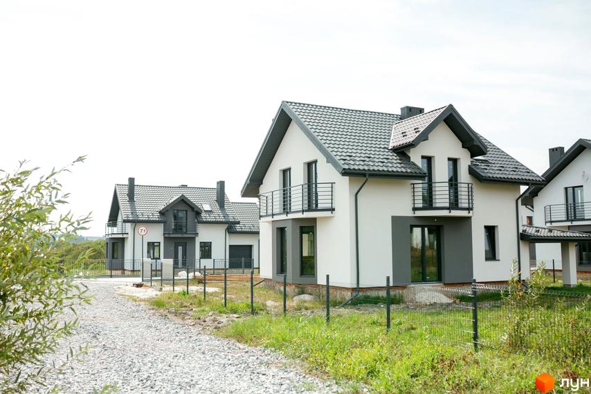 Котеджне містечко КМ Praud Hills з білими фасадами та сірими дахами. Навколо будинків зелена трава та гравійна дорога.