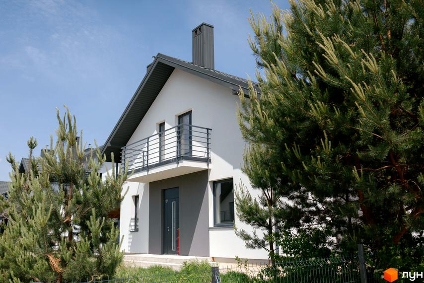 Котеджне містечко KM Praud Hills. Зовнішній вигляд білого будинку з сірим дахом та балконом, оточений зеленню.