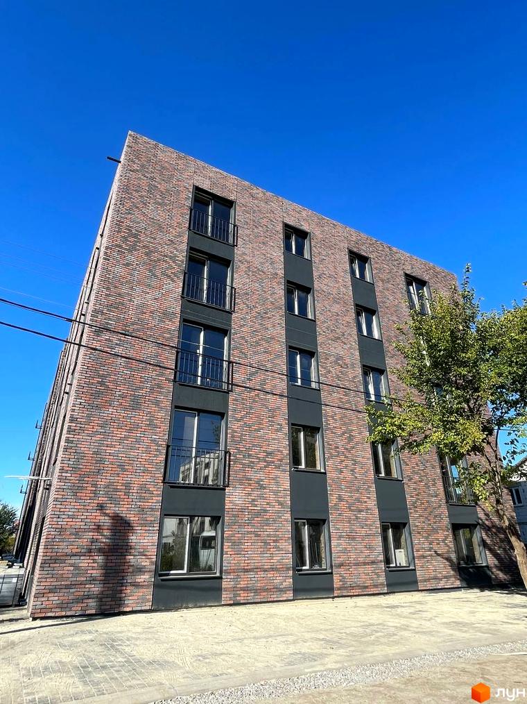 Фасад житлового комплексу Інститутська, оздоблений цеглою та темними панелями, з балконами та вікнами. Поруч розташована прибудинкова територія з бруківкою та деревом.