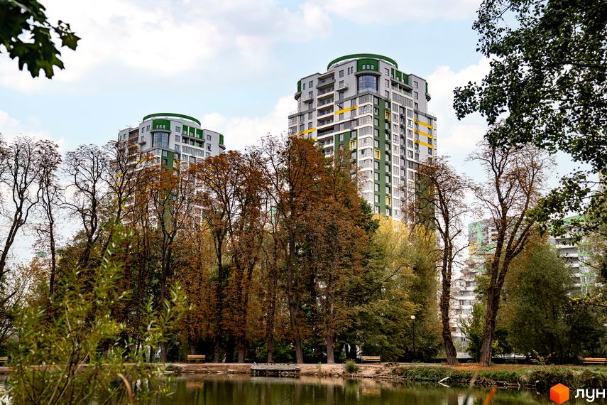 Житловий комплекс Паркове місто: високі будинки з сіро-зеленими фасадами та жовтими акцентами, розташовані біля парку з озером та осінніми деревами.