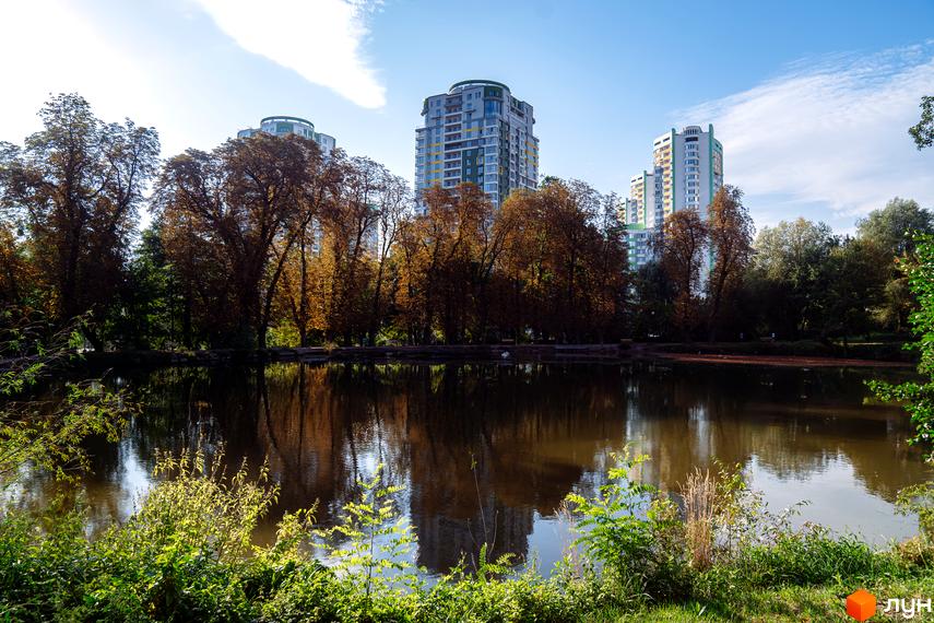 Багатоповерхові будинки Житлового комплексу Паркове місто з світлими фасадами та зеленими елементами. Перед ними озеро та осінні дерева, що відбиваються у воді.