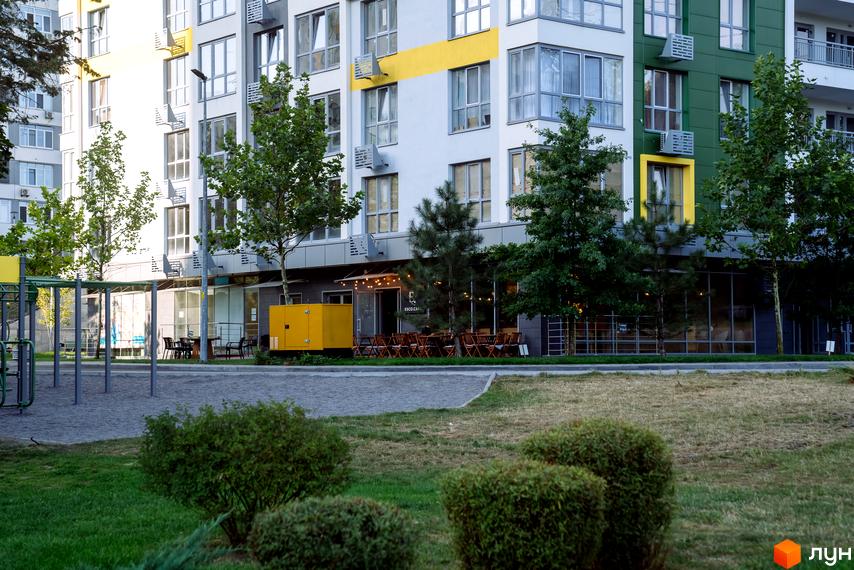 Багатоповерховий будинок Житлового комплексу Паркове місто з білими, жовтими та зеленими фасадами. Перед ним зелені насадження, дитячий майданчик та кафе.