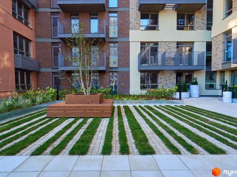 Фасад Будинку на Вавилових з цегли та світлих панелей, з балконами. Прибудинкова територія має дерев'яну лавку з деревом та доріжки з газону і плитки.