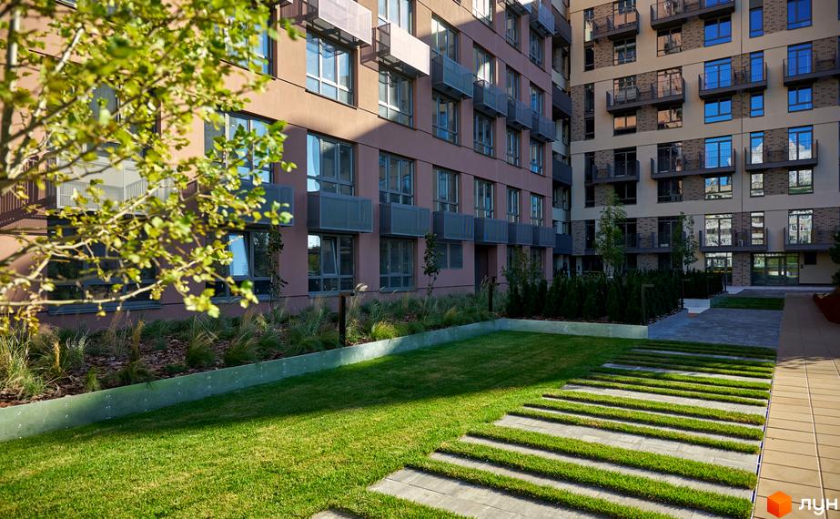 Багатоповерховий будинок житлового комплексу з терасами та зеленим газоном. Доглянута прибудинкова територія з доріжками та рослинами.