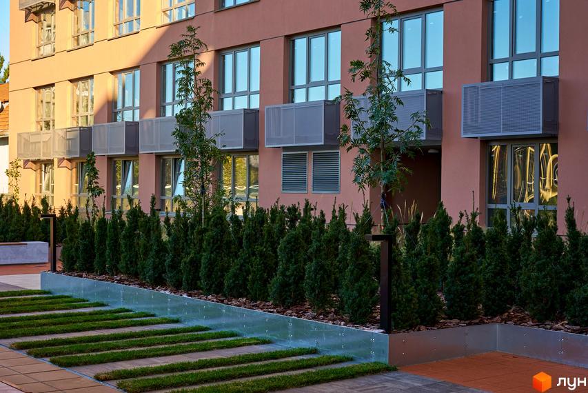 Будинок на Вавилових з теракотовим фасадом, великими вікнами та сірими захисними коробами. Прибудинкова територія має доріжки з трави, ряди кущів та молоді дерева.