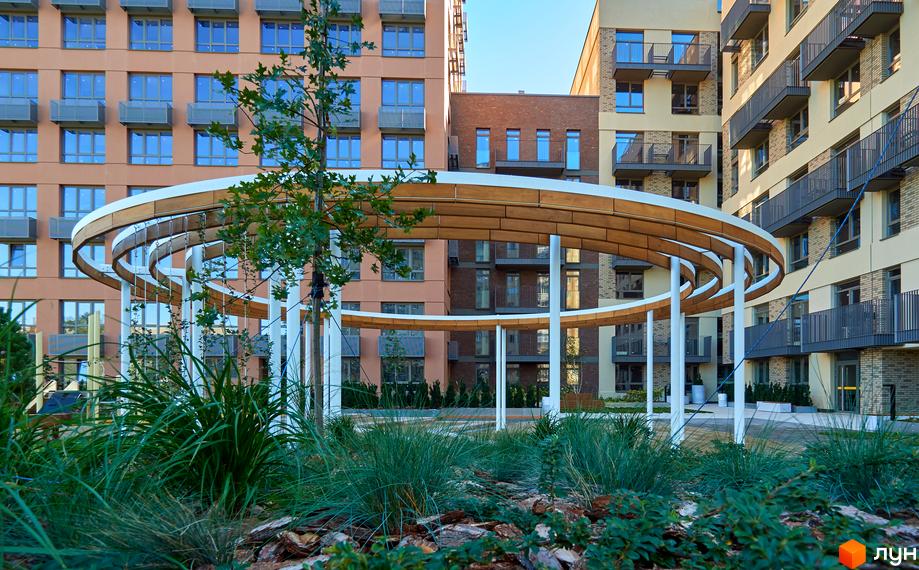 Житловий комплекс з фасадами пісочного та теракотового кольорів. На передньому плані - декоративна конструкція та зелені насадження.