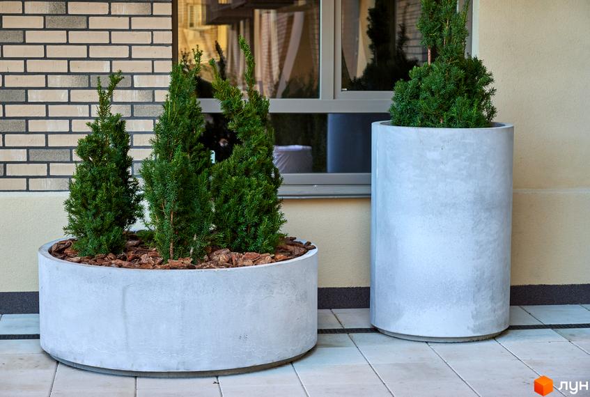 Фасад будівлі з цегляною кладкою та вікнами, поруч бетонні вазони з декоративними деревами. Благоустрій території.