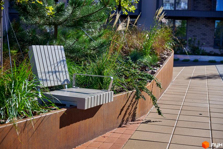 Фасад житлового будинку з панорамними вікнами. На прибудинковій території розташовані лавки для відпочинку та зелені насадження.