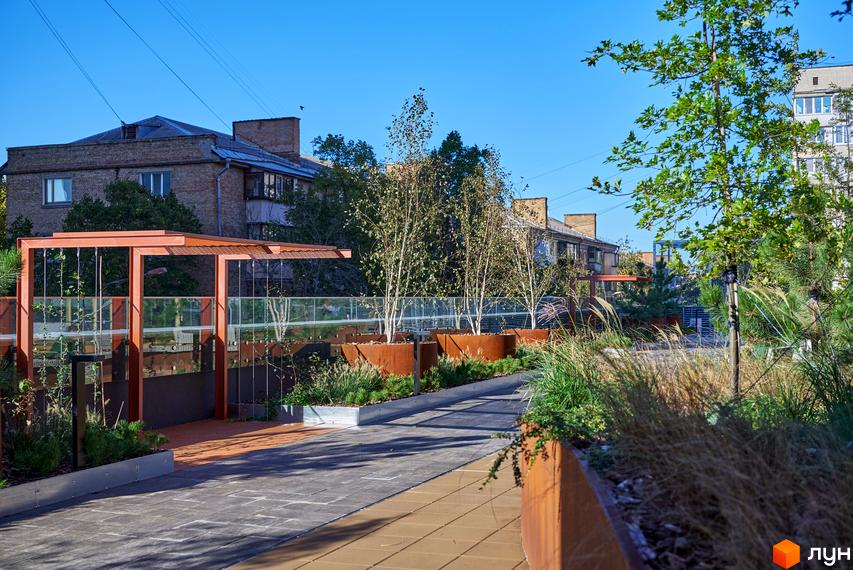 Будинок на Вавилових: багатоповерховий цегляний будинок, перед яким розташована доглянута прибудинкова територія з доріжками, металевими клумбами та різноманітними рослинами.