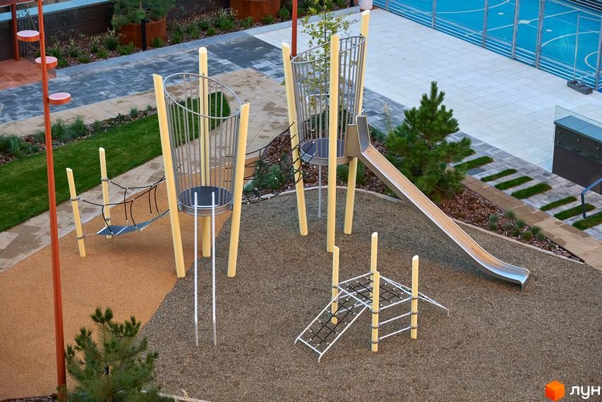 Ігровий майданчик у дворі ЖК Будинок на Вавилових з гіркою та мотузковим мостом для дітей, поруч зелені насадження та бруківка.