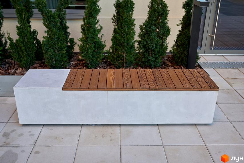 Бетонна лава з дерев'яним сидінням на тлі зелених рослин. Плитка на тротуарі перед будівлею.