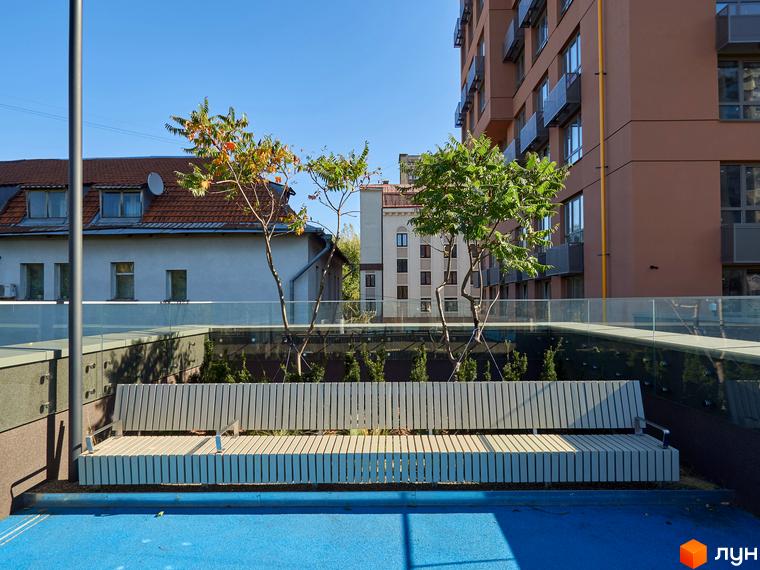 Фасад житлового комплексу Будинок на Вавилових з коричневими стінами та скляними балконами, поруч облаштована територія з лавкою та деревами.