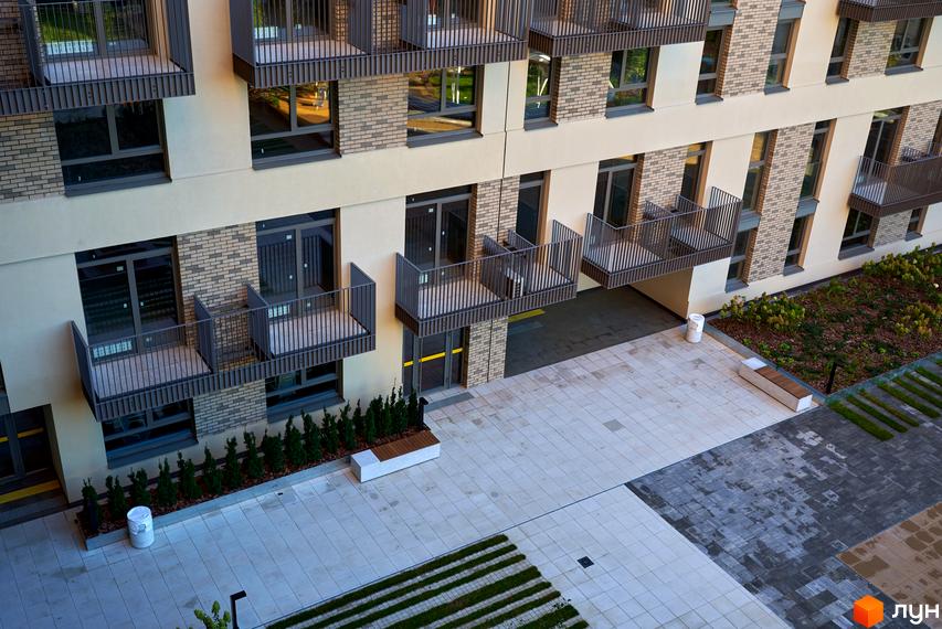 Фасад Будинку на Вавилових з бежевими стінами та цегляними вставками, оснащений балконами з темними поручнями. Прибудинкова територія має світлу плитку, зелені насадження та лавки.