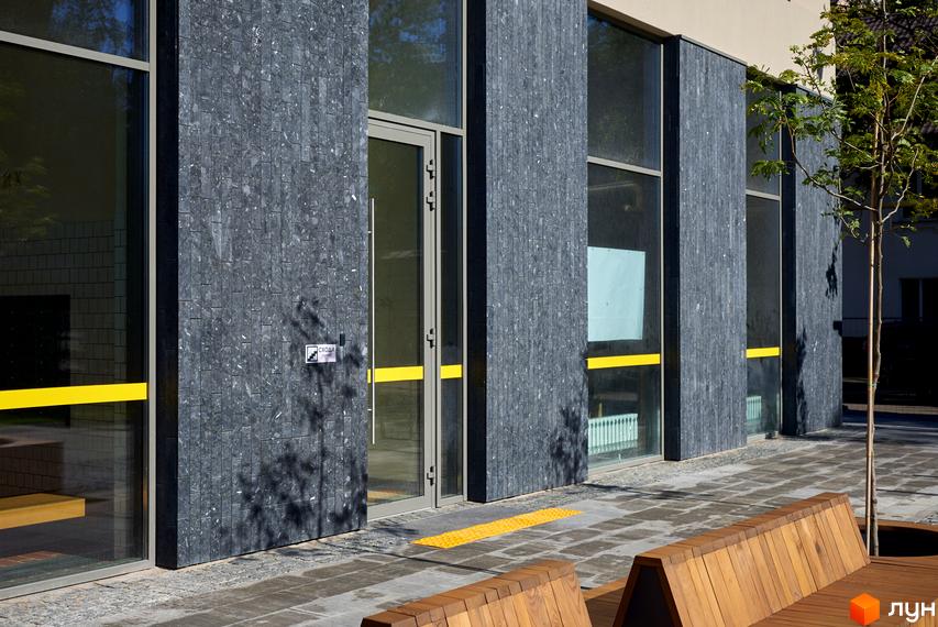 Фасад Будинку на Вавилових облицьований темно-сірою плиткою з великими вікнами. Прибудинкова територія облаштована дерев'яними лавками та деревами.