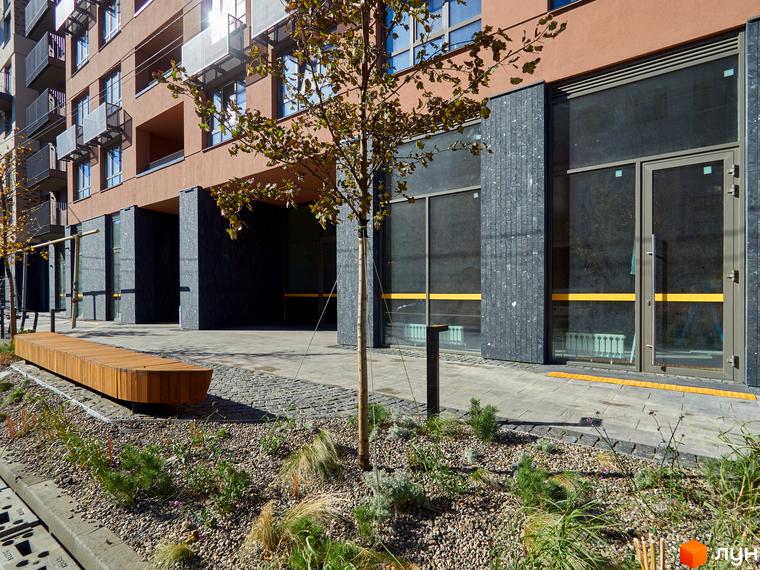 Житловий комплекс на Вавилових з фасадом теракотового кольору та темним цоколем. Прибудинкова територія має дерев'яну лавку та зелені насадження.