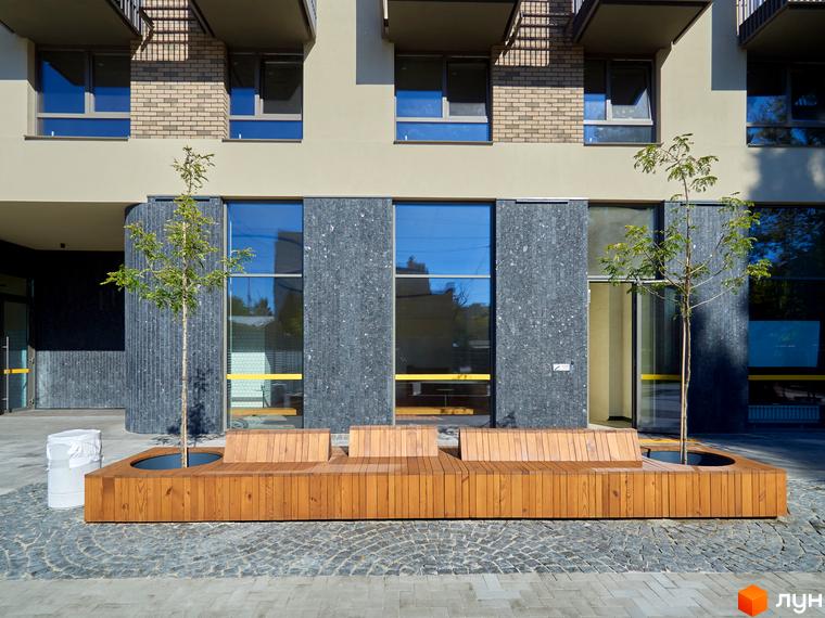 Фасад будинку ЖК Будинок на Вавилових з великими вікнами та декоративним оздобленням. На прибудинковій території розташовані лави та дерева.