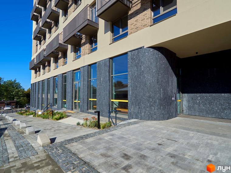 Зовнішній вигляд будинку на Вавилових з фасадом з темного каменю та скляними вікнами. На передньому плані бруківка та зелені насадження.