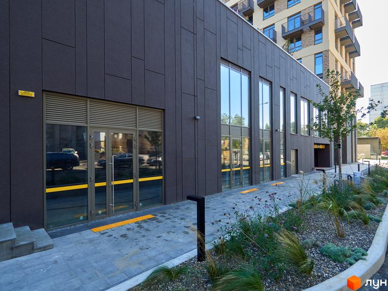 Темно-сірий фасад Будинку на Вавилових з великими вікнами та скляними дверима. Прибудинкова територія має доріжку з тактильною плиткою та озеленення.